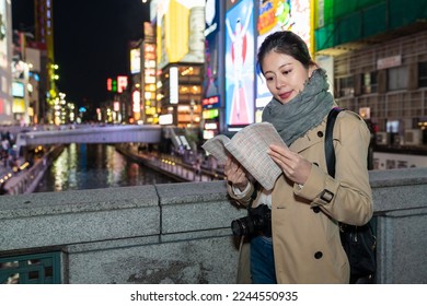 cheerful Asian Japanese female traveler reading guidebook on a bridge over doutonbori river at night in Shinsaibashisuji shopping area in Osaka japan during witner season - Shutterstock ID 2244550935