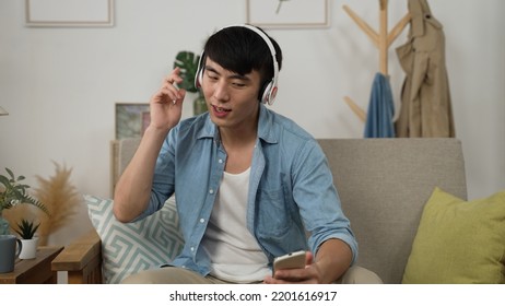 Cheerful Asian Japanese Young Guy With A Headset Is Bobbing Head To Rhythm After Choosing And Playing Songs On The Smartphone In The Living Room At Home