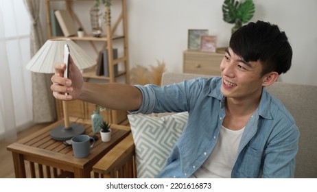 Cheerful Asian Japanese Young Male Is Waving Hi And Finger Pointing To The Mobile Phone Screen While Having A Video Chat With Friends During Quarantine On Sofa At Home.