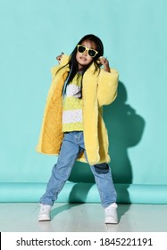 Cheerful Asian Kid Girl In Trendy Yellow Furry Coat, Blue Jeans, White Sneakers Is Fashion Posing Dancing With Her Hands Up Over Blue Wall Background. Trendy Children Fashion, Stylish Outfit Concept