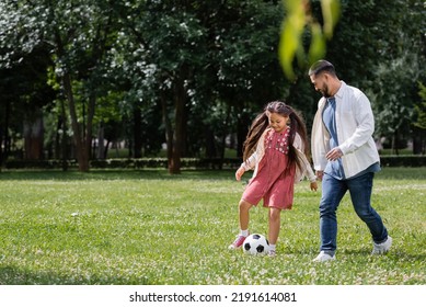 Cheerful Asian Kid Playing Football With Dad On Meadow In Park