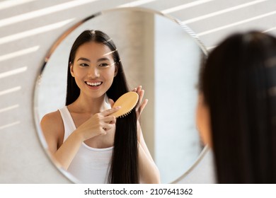 Cheerful asian lady with beautiful long hair standing in front of mirror at bathroom and combing her hair with wooden brush, smiling, closeup shot. Haircare Cosmetics Advert. Female Beauty Routine