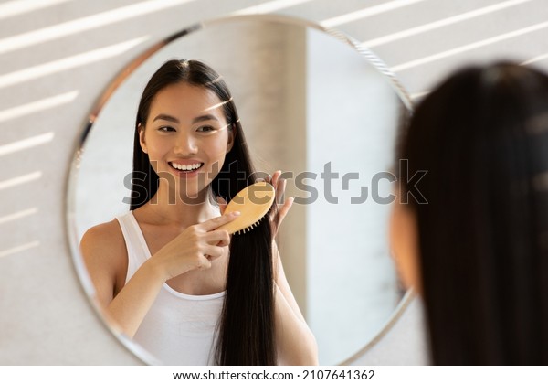 Cheerful asian lady with beautiful long hair\
standing in front of mirror at bathroom and combing her hair with\
wooden brush, smiling, closeup shot. Haircare Cosmetics Advert.\
Female Beauty Routine