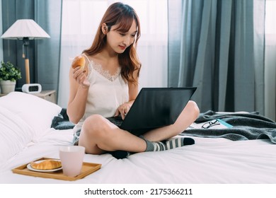 Cheerful Asian Lady Sitting Cross Legged On Bed Is Having Bread For Breakfast While Enjoying Social Media Feeds On The Computer At Leisure In A Bright Bedroom.