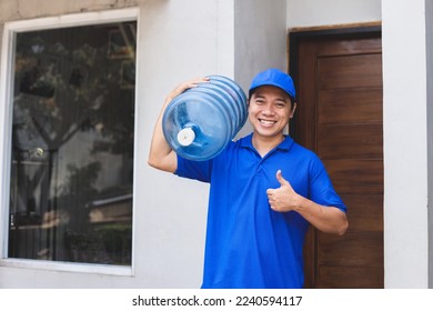 Cheerful Asian male courier in blue uniform, carry clear water gallon on shoulder, sending order to client and showing thumbs-up.