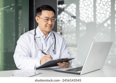 Cheerful Asian male doctor in a white coat works on his laptop and writes notes in a modern clinic office. Professional healthcare and medical consultation concept. - Powered by Shutterstock