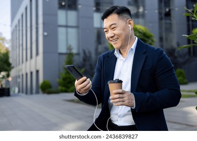 Cheerful Asian male executive enjoying a coffee break outside modern office buildings, using his phone with earphones. - Powered by Shutterstock