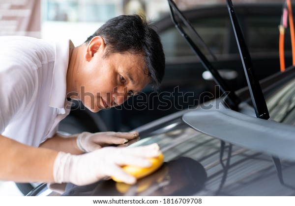 Cheerful Asian man cleaning and rubbing a car\
exterior by using sponge. Professional worker polishing a car with\
car wax, auto care\
concept.