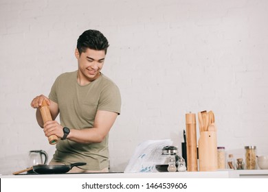 Cheerful Asian Man Cooking Breakfast While Reading Newspaper In Kitchen