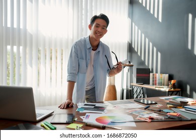 Cheerful asian man holding eyeglasses and smiling at camera while leaning on a table with a lot of creative stuff on it. Young designer in casual wear working in the modern office - Powered by Shutterstock