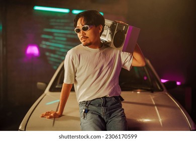 A cheerful Asian man is sitting on the hood of a vintage car outdoors and listening to music on an boombox. - Powered by Shutterstock