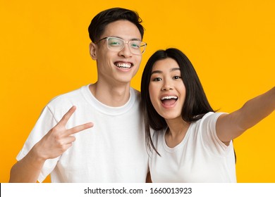 Cheerful Asian Man And Woman Taking Selfie, Man Showing Peace Sign, Yellow Studio Background