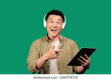 Cheerful asian middle aged man using digital pad and wireless headphones, listening to music on green background, smiling at camera and pointing at device - Powered by Shutterstock