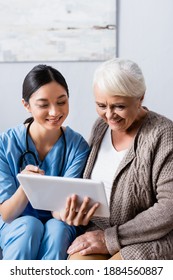 Cheerful Asian Nurse Pointing With Finger At Digital Tablet Near Smiling Senior Woman