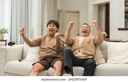 Cheerful Asian old man and middle aged son enjoy watching tv, celebrating good news. Smiling two male generations family feeling excited together, raising hands, resting on sofa in living room at home - Powered by Shutterstock