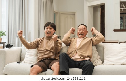 Cheerful Asian old man and middle aged son enjoy watching tv, celebrating good news. Smiling two male generations family feeling excited together, raising hands, resting on sofa in living room at home - Powered by Shutterstock