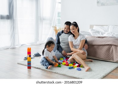 Cheerful Asian Parents Looking At Toddler Daughter Playing Building Blocks