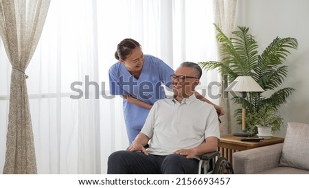 cheerful asian resident care attendant smiling while chatting with older man on wheelchair and encouraging him near the window at home during the day