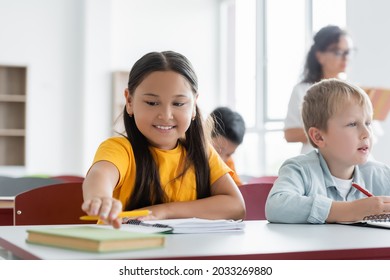 Cheerful Asian Schoolgirl Reaching Book While Sitting At Desk Near Classmate