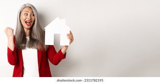 Cheerful asian senior woman buying house, scream of joy and making fist pump while showing paper house cutout, standing over white background. - Shutterstock ID 2311782195