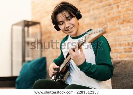 Cheerful Asian Student Guy Playing Electric Guitar Wearing Earphones Sitting On Sofa In Modern Living Room At Home. Boy Learning New Chords On Weekend. Hobby And Musical Gadgets. Selective Focus