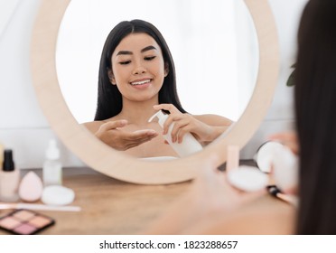 Cheerful asian woman cleaning her face, using cotton pads and cleansing product, sitting in front of mirror at home. Young smiling pretty lady applying cleansing milk on cotton pad, home interior