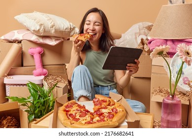 Cheerful Asian woman enjoys eating pizza and watching film on portable tablet poses on floor around cardboard boxes rents new apartment. Relocation at house. Tenancy and real estate concept. - Shutterstock ID 2176682641