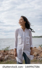 Cheerful Asian Woman With Happy Emotional Face And Beautiful Smile Standing Near River. Authentic Portrait Of Cute Korean Girl Walking, Enjoying Sea Landscapes, Breathing Fresh Air. Positive Lifestyle