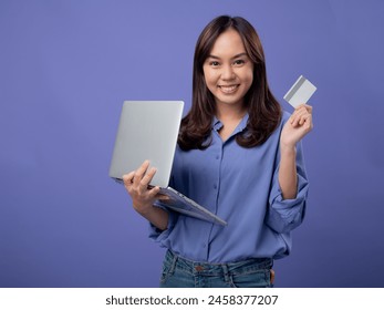 Cheerful Asian woman holding a laptop and a credit card, ready for online shopping, against a purple background - Powered by Shutterstock