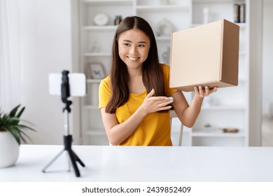 Cheerful asian woman influencer presenting package, preparing for an unboxing video, smiling young korean female recording with smartphone on tripod in bright, stylish room interior - Powered by Shutterstock