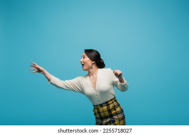 cheerful asian woman in plaid skirt smiling while gesturing isolated on blue