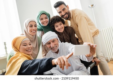 cheerful asian woman pointing with finger near muslim man taking selfie with interracial family