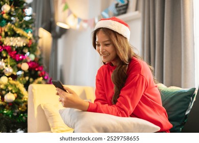 Cheerful Asian woman in red sweater and Santa hat seated on couch, smiling uses smartphone. Beautifully decorated Christmas tree stands in background, adding festive touch to cozy - Powered by Shutterstock