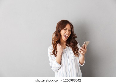 Cheerful asian woman standing isolated over gray background, holding mobile phone, celebrating