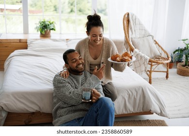 Cheerful asian woman in warm clothes holding sweet pastry near african american boyfriend with coffee in bedroom - Powered by Shutterstock