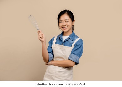 Cheerful Asian woman wearing apron and holding hand whisk standing isolated on light brown background. - Powered by Shutterstock