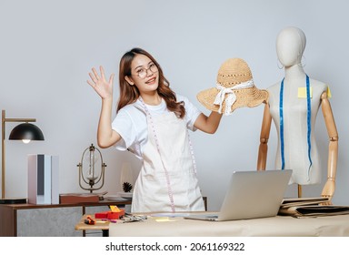 Cheerful Asian Young Female Designer Wearing Eyeglasses Doing Fashion Design On Beautiful Sombrero In Studio, Full Of Tailoring Tools  With Mannequin And Equipment On The Desk