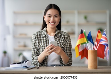 Cheerful Asian Young Lady Using Smartphone, Having English Or Other Foreign Language Class, Sitting At Table With Wireless Headset And International Flags At Home, Using Mobile Application, Copy Space