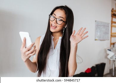 Cheerful Asian Young Woman In Glasses And Earphones Making Video Call Using Mobile Phone