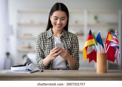 Cheerful Asian Young Woman Using Cellphone, Having English Or Other Foreign Language Class, Sitting At Table With Wireless Headset And International Flags At Home, Using Mobile Application, Copy Space