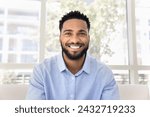 Cheerful attractive African freelance business man talking on video conference call from home, sitting on couch, looking at camera with toothy smile. Screen view head shot portrait