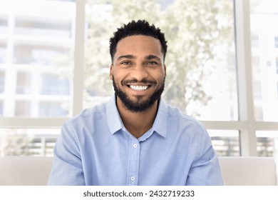 Cheerful attractive African freelance business man talking on video conference call from home, sitting on couch, looking at camera with toothy smile. Screen view head shot portrait