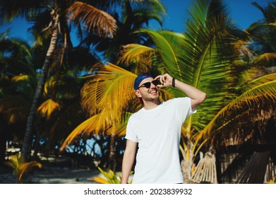 Cheerful attractive bearded tourist boy in white tshirt smiling broadly while adjusting sunglasses against palm trees - Powered by Shutterstock