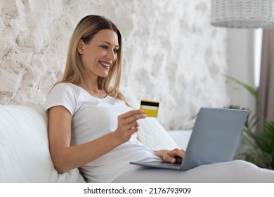 Cheerful Attractive Blonde Woman In Pajamas Sitting In Bed With Brand New Computer And Yellow Plastic Credit Card, Enjoying Weekend At Home, Order Food Online, Side View, Copy Space