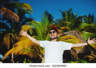 Cheerful attractive boy in white t shirt and sunglasses, gesturing triumph with raised hands against palm trees background