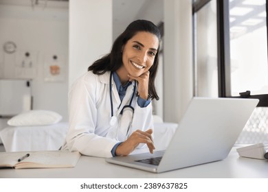 Cheerful attractive doctor woman professional portrait. Young Latin practitioner sitting at workplace table with laptop, looking at camera with toothy smile posing for shooting in surgery office - Powered by Shutterstock