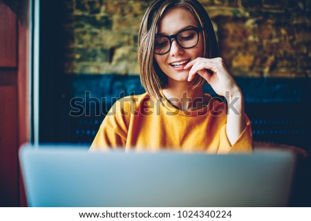 Cheerful attractive female student with eyeglasses and short haircut chatting online in social networks with friend on modern computer using free high speed internet connection sitting in coffee shop