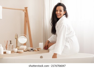 Cheerful Attractive Lady Wearing Bathrobe Sitting On Bathtub Smiling To Camera Posing In Modern Bathroom At Home. Wellness And Spa, Beauty Routine Concept