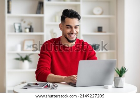Cheerful attractive millennial arabic businessman with beard in red clothes typing on laptop in home office interior. Education, work remote, blog and modern technology during covid-19 pandemic