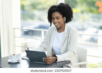 Cheerful attractive millennial black woman in formal outfit sitting at table at terrace, drinking coffee, have video call with business partner, using digital tablet, cafe interior, copy space - Powered by Shutterstock